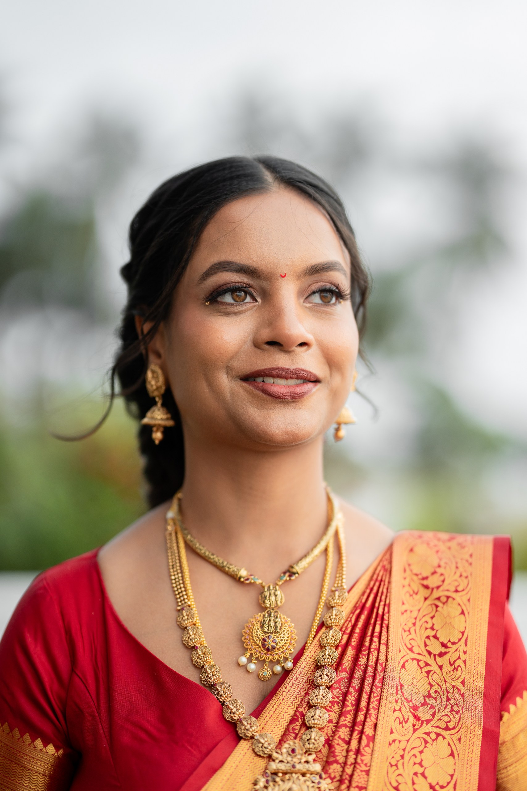 A woman wearing traditional Indian attire, featuring a red saree with golden embroidery and ornate jewelry, including a necklace and earrings. She has a confident smile and a bindi on her forehead, set against a blurred natural background.